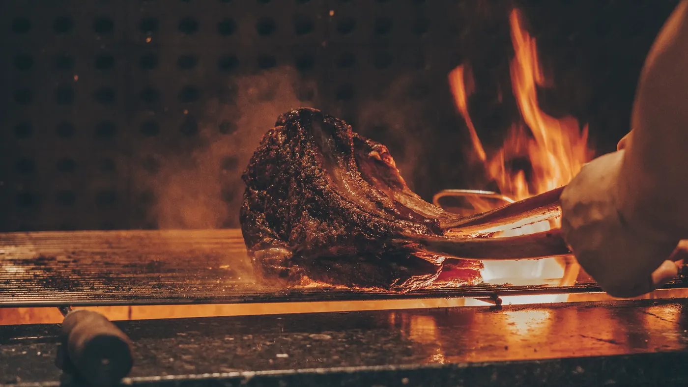 Kotlet wołowy z kością zdejmowany z rusztu, za nim płomienie z drewna bukowego