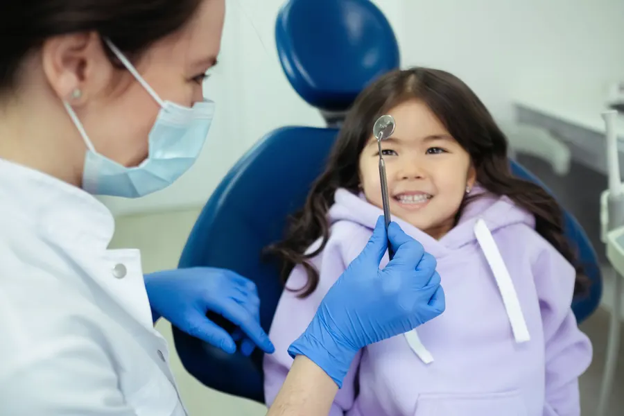 Uśmiechnięta dziewczynka na fotelu dentystycznym, obok lekarka z lusterkiem